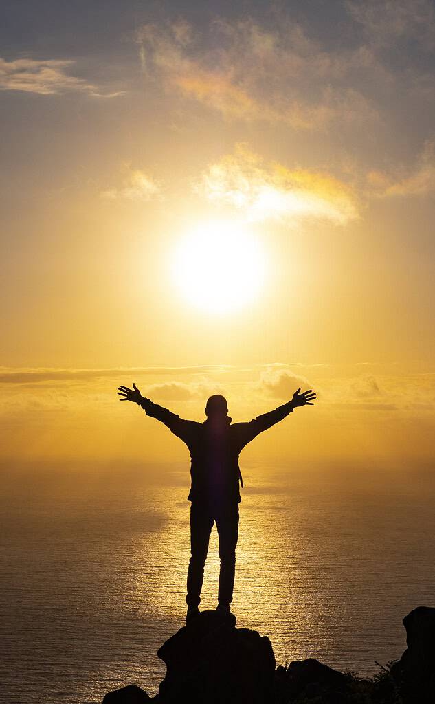 Person standing with arms raised toward the sun, silhouetted at sunrise